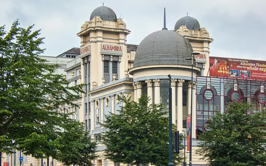City centre street in Bradford featuring a red - brick Victorian building, a traditional fish and chips shop, and ornate stone buildings, with trees and people walking in the square.